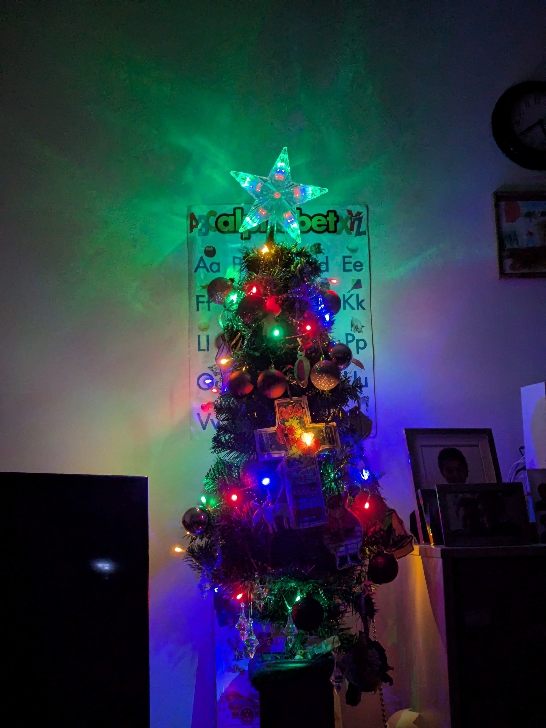 Auto-generated description: A decorated Christmas tree is illuminated with colorful lights and topped with a glowing star, set against a background that includes an alphabet chart and a few framed photos.