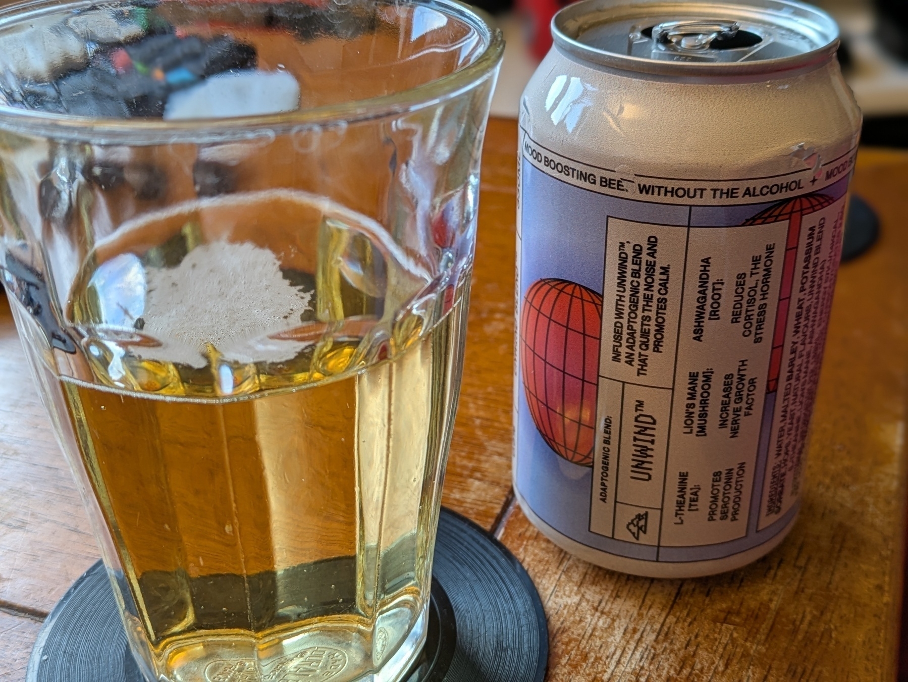 A glass with a pale yellow beverage sits on a coaster next to a can of non-alcoholic beer on a wooden table.