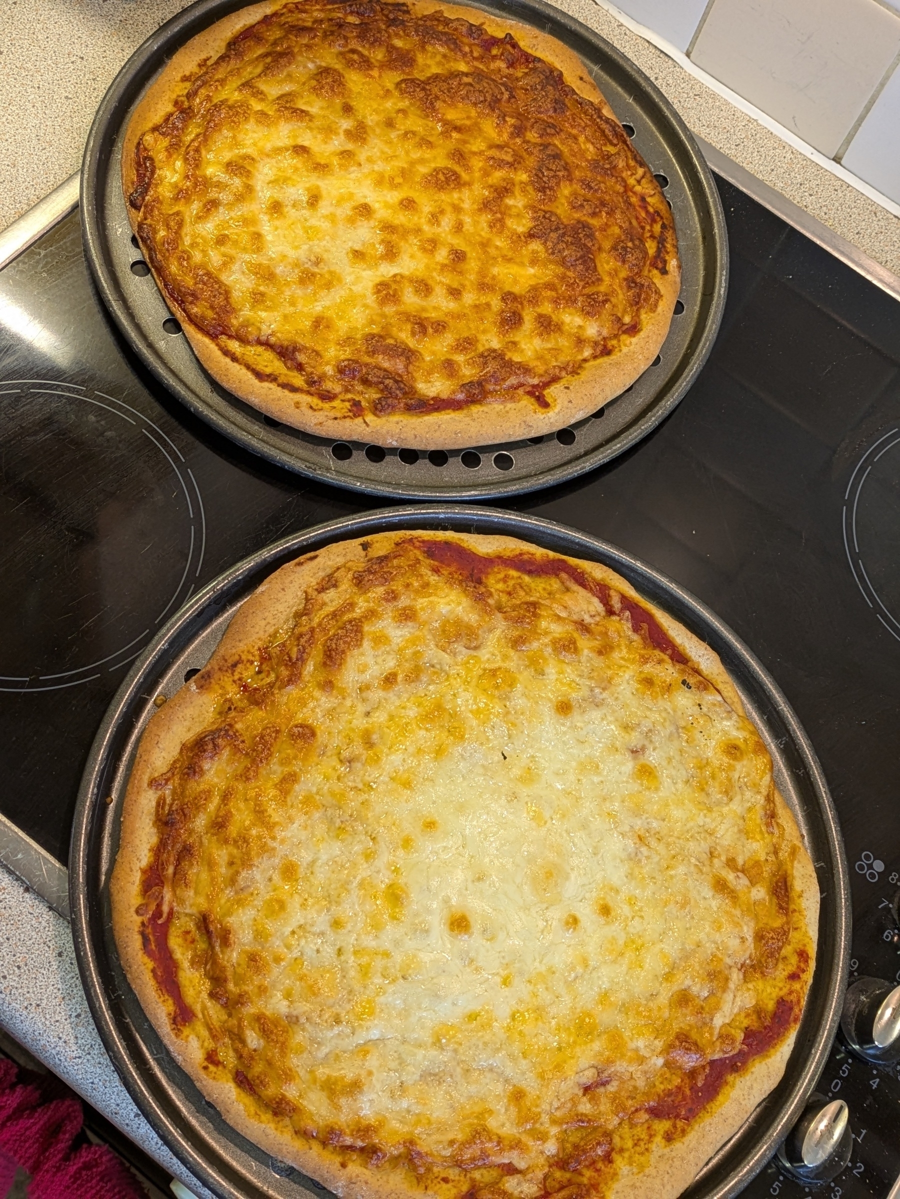 Two freshly baked cheese pizzas are cooling on round trays on a stovetop.